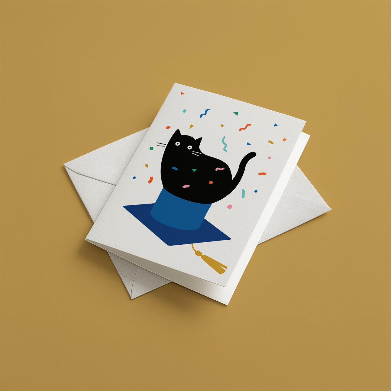 Greeting card with a black cat design on a white background, featuring a blue cape and colorful confetti, with an envelope on a brown background.