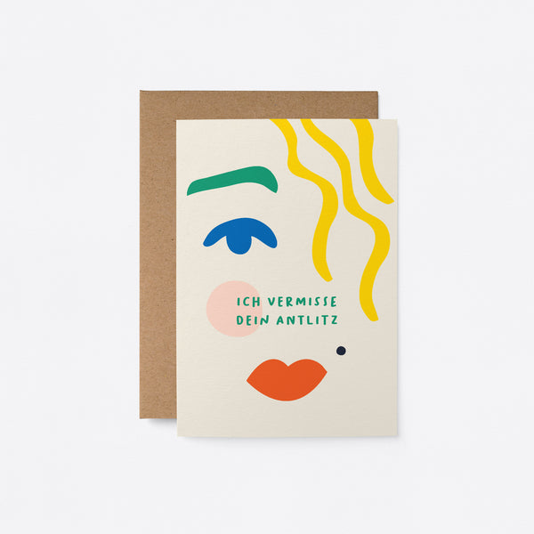 German Greeting card with blue eye, green brow, red lips, yellow hair and a text that says Ich vermisse dein Antlitz