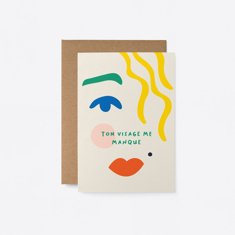 French Greeting card with blue eye, green brow, red lips, yellow hair and a text that says Ton visage me manque