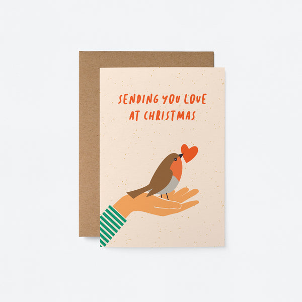Christmas card with a bird and a red heart in its beak on the palm of a hand and a text that says sending you love at christmas