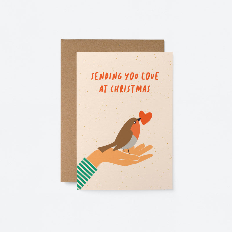 Christmas card with a bird and a red heart in its beak on the palm of a hand and a text that says sending you love at christmas