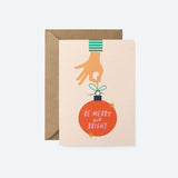 christmas card with a hand holding red christmas tree ornament and a text in it that says be merry and bright